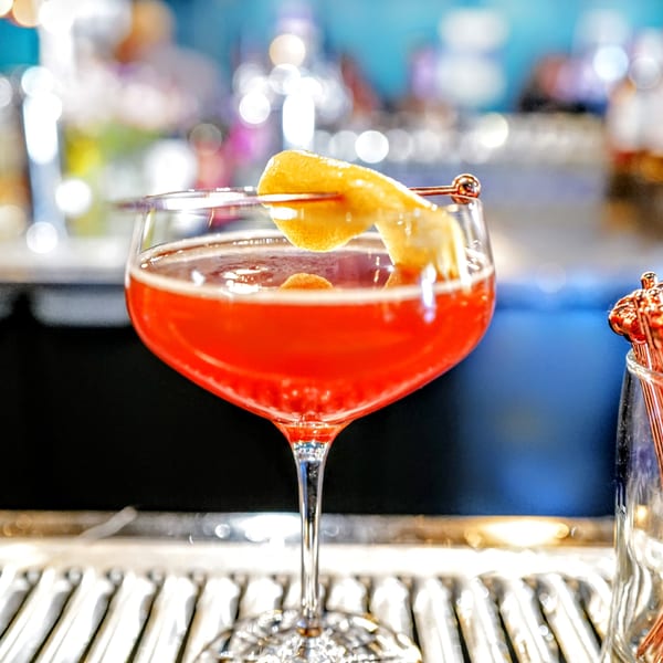 A red cocktail in a stemmed glass, garnished with a lemon peel, on a bar.