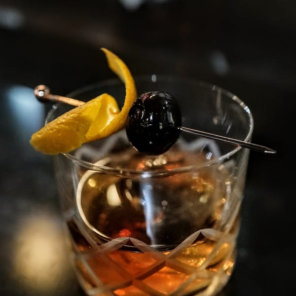 Close-up of an Old Fashioned cocktail with an orange peel and dark cherry garnish.
