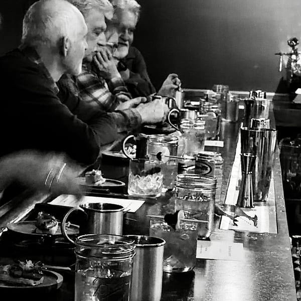 Black and white image of three men sitting at a bar with drinks and small plates.
