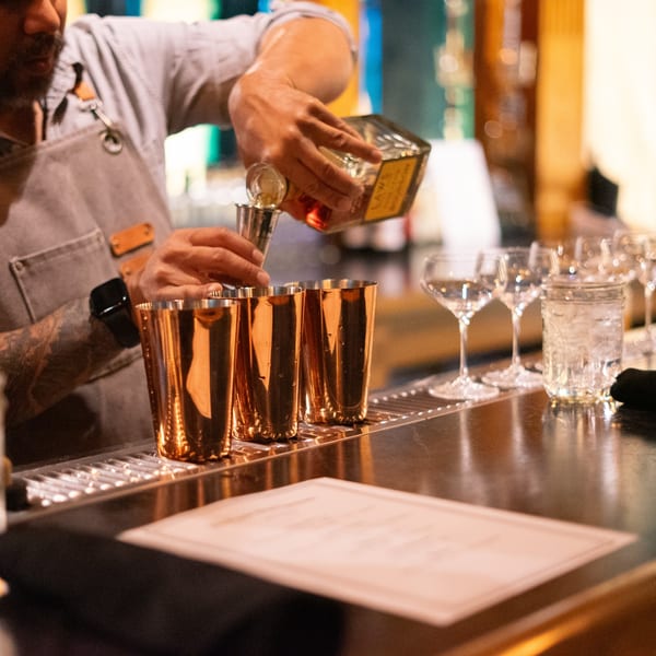 Bartender pouring liquor from a bottle into a jigger over copper cocktail shakers on a bar.