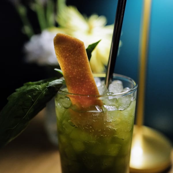 Green cocktail with ice, an orange peel garnish, and a dark straw, against a blue background.