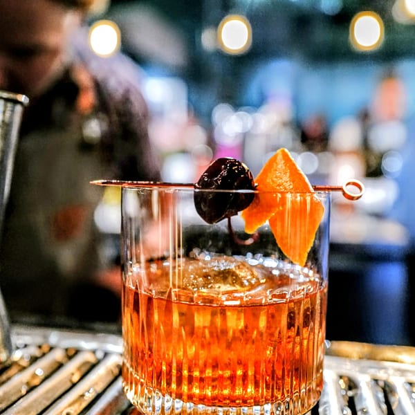 An Old Fashioned cocktail with an orange peel and cherry garnish, on a bar.