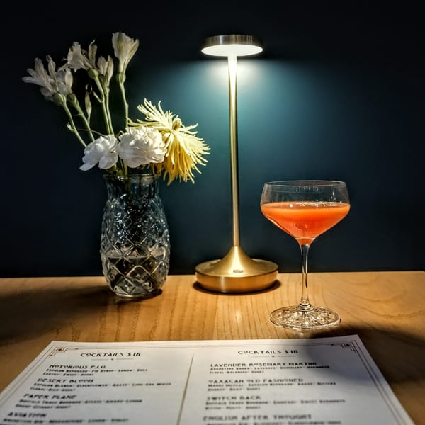 A cocktail, menu, flowers, and lamp on a wooden table against a dark blue wall.
