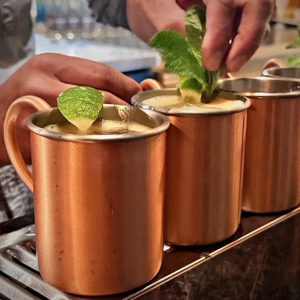 Bartender garnishing three copper mugs filled with Moscow Mules with fresh mint and lime.