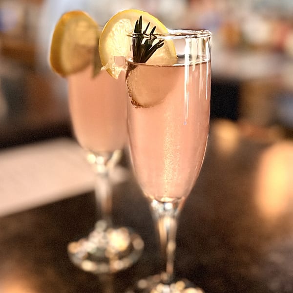 Two champagne flutes filled with pink cocktails, garnished with lemon slices and rosemary sprigs.