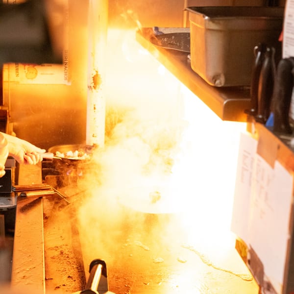 A chef's hand cooking food on a fiery grill, with bright orange flames and smoke.