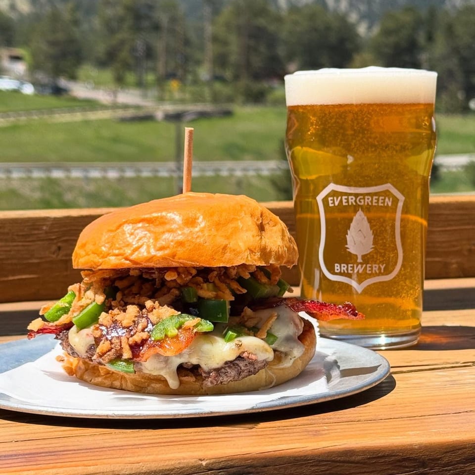 A burger with bacon, jalapeños, and crispy onions next to an Evergreen Brewery beer outdoors.