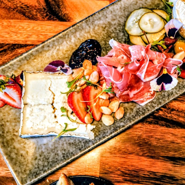 Charcuterie board with cheese, prosciutto, strawberries, almonds, fig jam, and pickles on a wooden table.