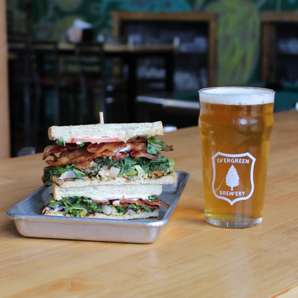 A tall club sandwich with bacon and greens next to a pint of Evergreen Brewery beer.